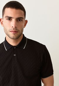 Zwart poloshirt met een gestructureerd chevronpatroon, voorzien van een contrasterende grijze kraag en knopen. Korte mouwen met grijze accenten.