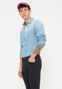 Lyseblå denimskjorte med rullede ermer, hvit undertrøye og svarte bukser. Har en subtil tekstur. Modellen står med hånden i lommen.