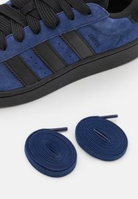 Blå ruskindsneakers med sorte striber og sorte snørebånd, ledsaget af to sammenrullede blå snørebånd på en hvid overflade.