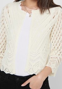 Lace-knit cardigan in cream, featuring a scalloped hem, three-quarter sleeves, and a lightweight, open pattern. Worn over a white top.