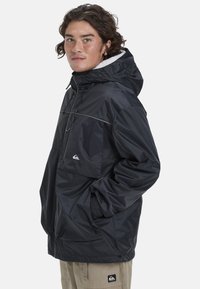 Black waterproof jacket with a hood, zipper pocket, half-zip front, and a logo on the chest. Features elastic cuffs and a loose fit.