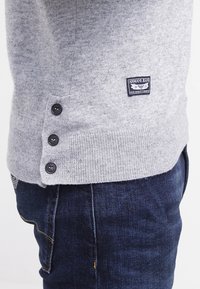 Ourlet de pull en maille gris avec trois boutons noirs et logo Armani Jeans au-dessus de la poche du jean en denim bleu foncé.
