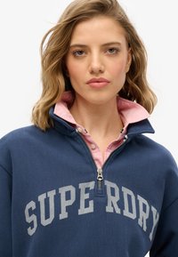 Granatowy polar z suwakiem z szarym napisem "SUPER DRY". Posiada różowy kołnierz, miękką teksturę oraz srebrny detal suwaka.