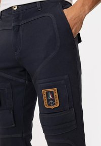 Pantaloni cargo della marina con molte tasche, cuciture in contrasto e un badge con un design tricolore sulla coscia.