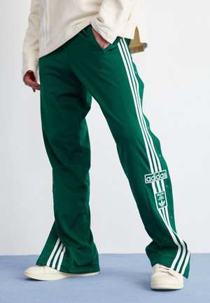 Person trägt grüne Adidas-Trainingshose mit weißen Streifen und Druckknöpfen an den Seiten, kombiniert mit weißen Sneakers und einem hellen Oberteil.