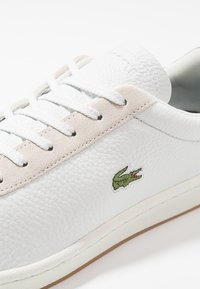 Baskets en cuir blanc avec une finition texturée, accents en daim beige, lacets blancs et un logo crocodile brodé vert sur le côté.