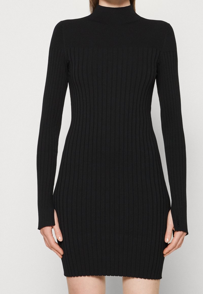 Robe noire en tricot côtelé avec un col montant, des manches longues et une coupe ajustée. Présente une surface texturée et un ourlet festonné.
