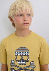 Gul bomulds T-shirt med en stor kraniegrafik med geometriske mønstre i blå, brun og hvid. Rund halsudskæring, korte ærmer.