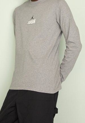 Personne portant un t-shirt Jordan gris à manches longues et un pantalon noir, debout avec une main derrière le dos sur un fond uni.