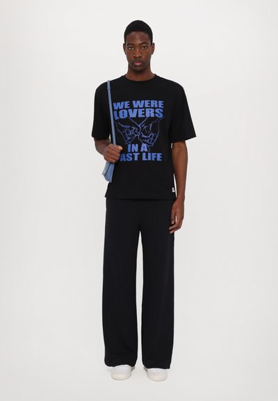 Black t-shirt with blue graphic text "WE WERE LOVERS IN A PAST LIFE," paired with loose dark trousers and white sneakers.