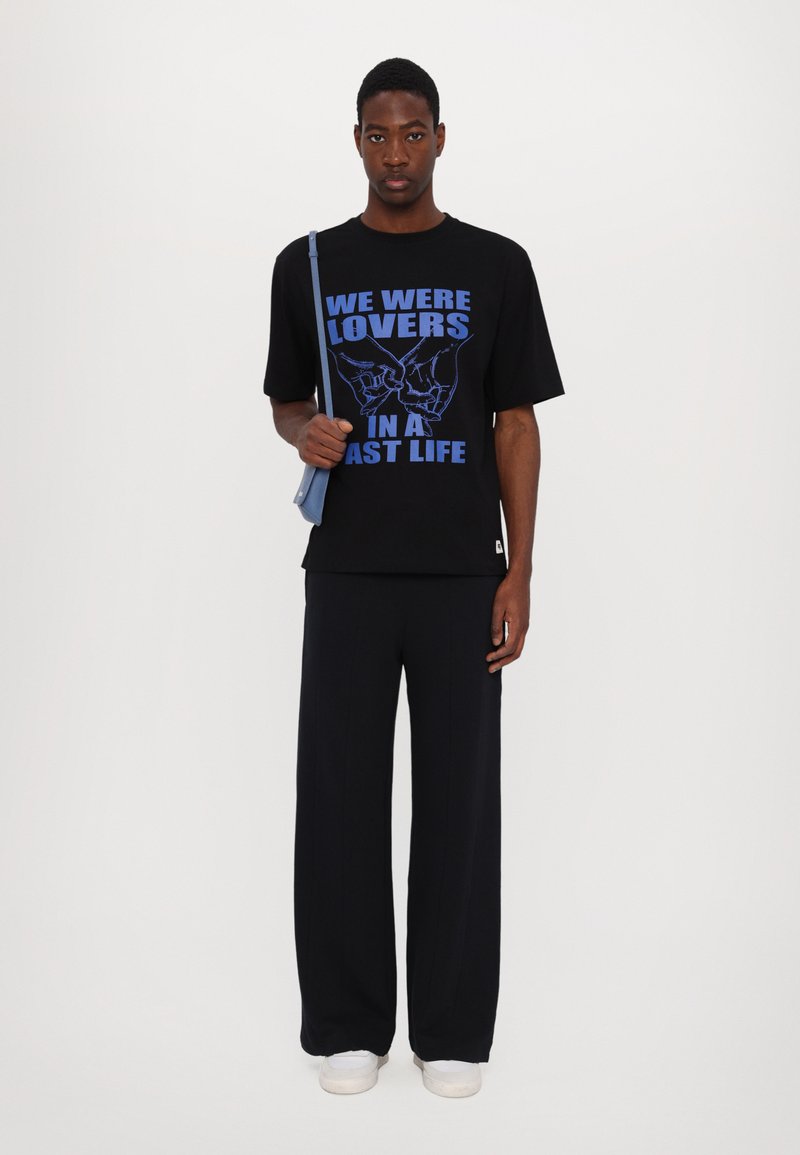 Schwarzes T-Shirt mit blauer Grafikschrift "WIR WAREN LIEBHABER IN EINEM FRÜHEREN LEBEN", kombiniert mit lockeren dunklen Hosen und weißen Turnschuhen.