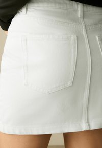 Jupe en denim blanc avec un design ajusté, des poches arrière et un ourlet propre. Le tissu a une texture douce avec une légère élasticité.