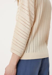 Beige pletený pulovr s tříčtvrtečními rukávy. Má strukturované pruhy a vyšívané detaily. Hladký pas a rukávy.