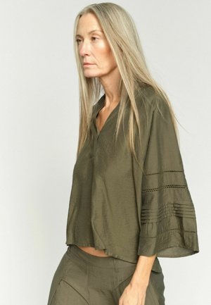 Žena s dlhými blond vlasmi, oblečená v voľnej olivovo zelenej blúzke a zodpovedajúcich nohaviciach, stojaca pred jednoduchým svetlým pozadím.