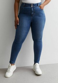 Vaqueros ajustados de cintura alta en denim azul oscuro. Cuenta con bolsillos delanteros y cierre de botones. Combinados con zapatillas blancas.
