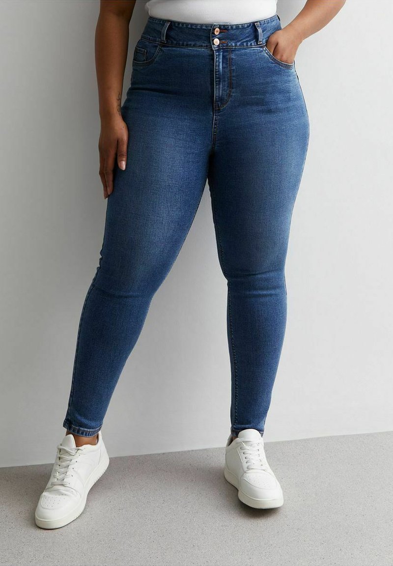 Vaqueros ajustados de cintura alta en denim azul oscuro. Cuenta con bolsillos delanteros y cierre de botones. Combinados con zapatillas blancas.