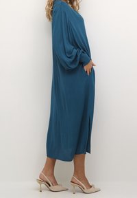 Tealfarbenes, langärmliges Kleid mit lockerer Passform, seitlichem Schlitz und strukturiertem Stoff. Kombiniert mit hellen, spitz zulaufenden Absätzen.