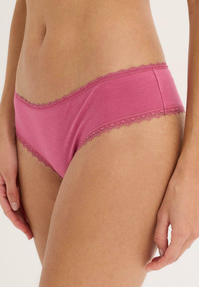 Femme portant un sous-vêtement hipster en coton avec des bordures en dentelle rose, montrant le torse, les hanches, et la main sur un fond uni.