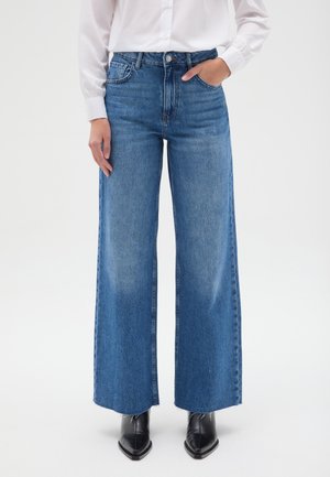 Persona que lleva vaqueros azules de tiro alto y pierna ancha, camisa blanca abotonada y botas negras de punta afilada, de pie con una mano en el bolsillo.