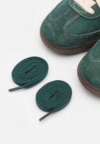 Gröna mockaskor med bruna sulor bredvid två prydligt ihoptrullade mörkgröna skosnören på en vit yta.