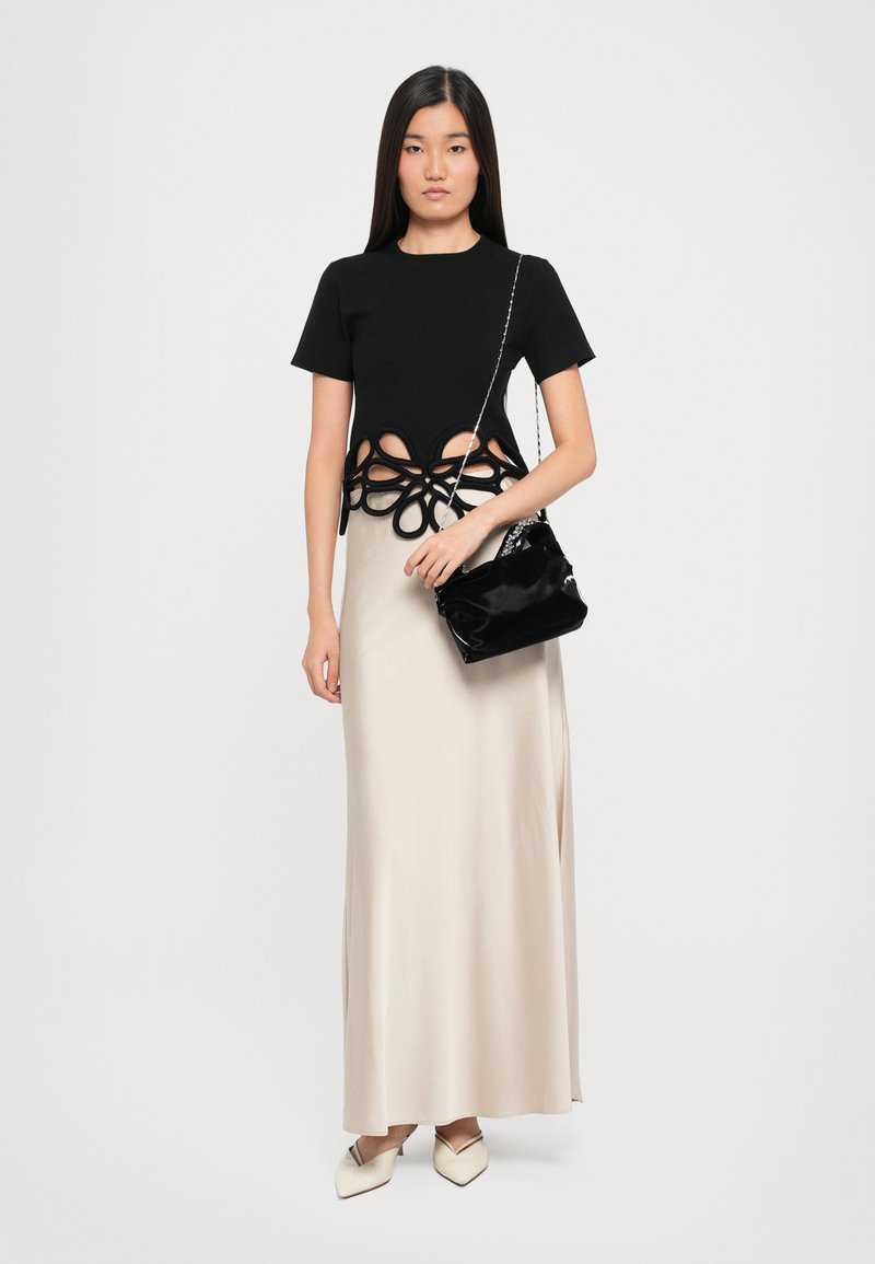 T-shirt noir court avec un motif floral découpé, associé à une longue jupe maxi en satin beige élégante. Le mannequin tient un petit sac à bandoulière noir.