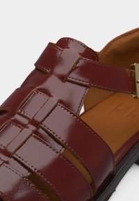 Καφέ λουστρινη σαγιονάρα ψαρά με ρυθμιζόμενο λουράκι με αγκράφα και μαύρη σόλα, δείχνοντας σχεδιασμό πλεγμένου λουριού και ταμπά εσωτερικό.