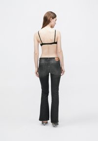 Čierne rozšírené džínsy s vyblednutou textúrou a viditeľným šitím; na zadnej strane je nášivka s menovkou značky. Modelka má na sebe čierny bikinkový top.