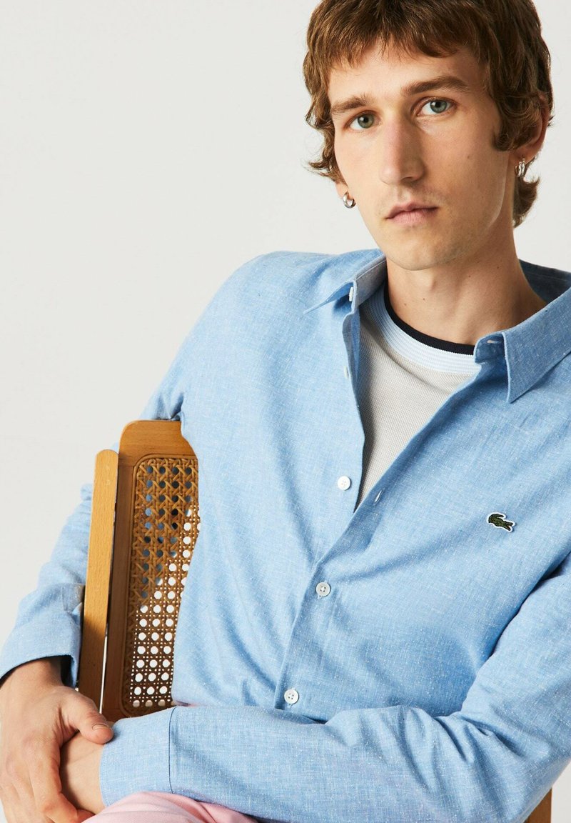 Junger Mann mit hellbraunem Haar, der ein hellblaues, geknöpftes Hemd mit einem kleinen Krokodil-Logo trägt und auf einem Holzstuhl mit geflochtener Rückenlehne sitzt.