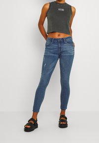 Top corto sin mangas gris con texto impreso, acompañado de jeans ajustados desgastados azules y sandalias negras con correas ajustables.