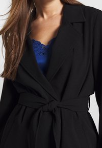 Mustaan bleiseri, jossa on sidottu vyötärö ja läpän kaulus. Alla näkyy syvänsininen paita, jossa on kukkakuvioinen pitsireunus.