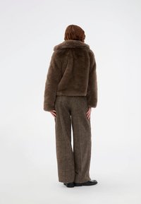 Brahna umetni krzneni jopič z širokim ovratnikom, v kombinaciji s širokimi hlačami v rjavem vzorcu in črnimi čevlji, s pogledom s hrbta.