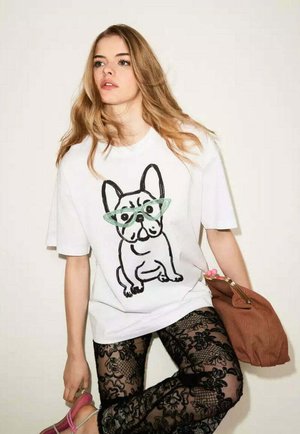 Giovane donna con lunghi capelli biondi che indossa una maglietta bianca oversize con un'illustrazione di cane e leggings in pizzo nero con motivo, tenendo una borsa marrone.