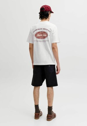 Joven con camiseta blanca con gráfico, pantalones cortos negros, zapatos marrones, calcetines oscuros y gorra roja, de pie de espaldas sobre un fondo liso.