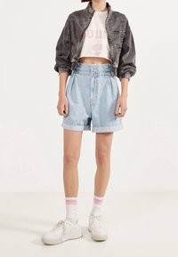 Ljusblå högmidjade denimshorts med upprullade fållar, tillsammans med en kort vit t-shirt med grafik och en mörkgrå bomberjacka. Vita sneakers.