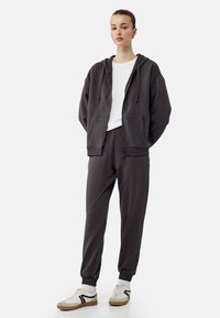 Mørkegrå zip-up hoodie med frontlommer, parret med matchende joggerbukser med elastiske manchetter. Hvid t-shirt med rund hals underneath.