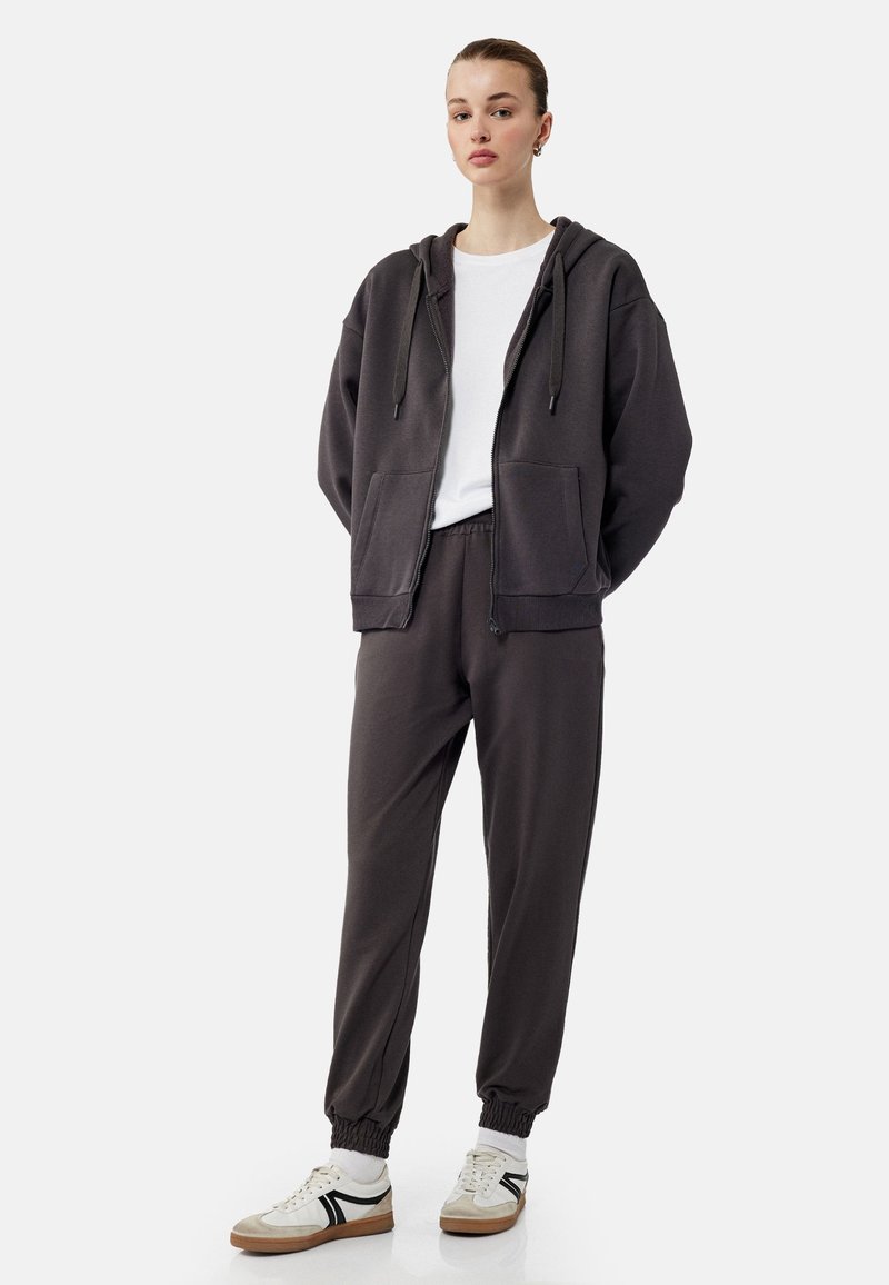 Mørkegrå zip-up hoodie med frontlommer, parret med matchende joggerbukser med elastiske manchetter. Hvid t-shirt med rund hals underneath.