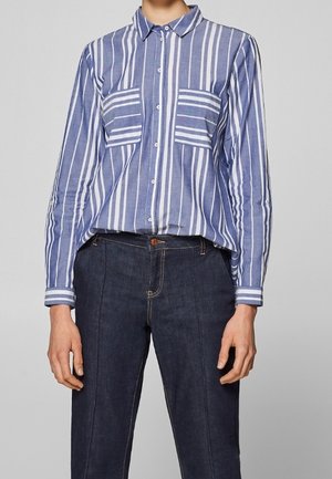 Blauw-wit gestreept overhemd met knopen, verticale strepen en twee horizontaal gestreepte borstzakken, gedragen met donkerblauwe hoge taille jeans.