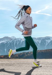 Lysegrå løbejakke med hætte, sorte detaljer; grønne leggings; hvide og sorte løbesko med gule detaljer; sneklædte bjerge i baggrunden.