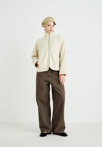 Woman wearing beige fleece zip-up jacket, beige knit beret, wide brown pants, and black flats, standing against a plain white background.