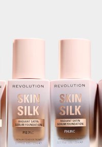 Revolution Skin Silk foundationflasker med jevn rosa emballasje og metallisk gulltekst, merket med nyansene F12.7NC og F14.9NC.