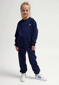 Mörkblå sweatshirt och matchande joggers i mjukt tyg. Sweatshirten har en liten hjärtlogotyp på bröstet. Vita sneakers med blå detaljer.