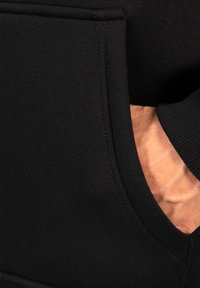 Sort sweatshirt med kængurulomme, glat struktur og ribbede manchetter. Nærbillede viser en hånd, der rækker ind i lommen, hvilket fremhæver sømmene.