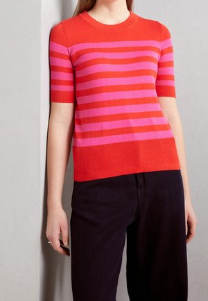 Pull court à manches courtes rayé rouge et rose avec un col rond, doté de poignets et d'un ourlet côtelés, associé à un pantalon sombre.