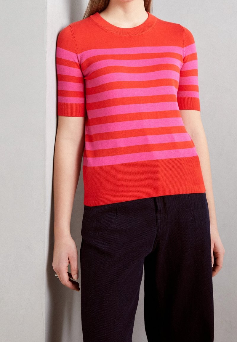 Punainen ja vaaleanpunainen raidoitettu lyhythihainen neulepaita, jossa on pyöreä kaula-aukko, ripustettuna ribbaavilla hihansuilla ja helmoilla, yhdistettynä tummiin housuihin.