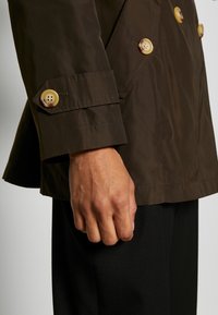 Höger hand på person som bär mörkbrun knapplöst jacka och svarta byxor mot en enkel ljus bakgrund.