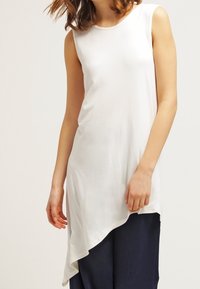 White sleeveless top with a round neckline and asymmetric hem, made of smooth fabric, paired with navy blue pants.