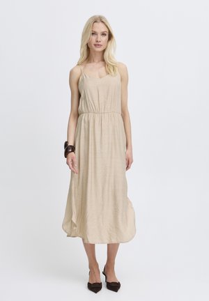 Lyshåret kvinde iført en beige ærmeløs midikjole med indsnøret talje, mørkt armbånd og mørke spidse hæle, stående mod en ensfarvet baggrund.