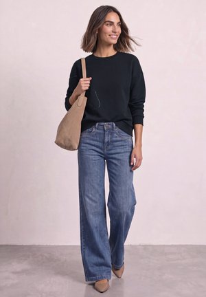 Femme portant un sweat-shirt noir et un jean large, avec un sac beige à l'épaule et souriante, debout sur un sol gris avec un fond lumineux.