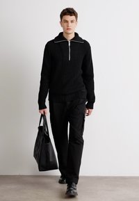 Suéter de punto canalé negro con cuello con cremallera, combinado con pantalones cargo negros. El modelo lleva un bolso tote negro texturizado y calza zapatos negros.