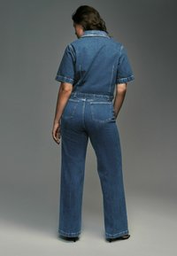 Bermuda en denim bleu marine, à manches courtes, au design à col, taille ajustée, pantalons larges, poches arrière, texture lisse et accents minimalistes.